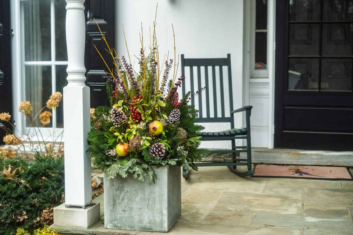 Decorative porch scene with a concrete planter filled with winter foliage, pinecones, and ornaments beside a dark green rocking chair on a stone patio.