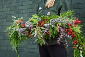 A person in a green jacket holds a festive garland with pine branches, red berries, silver pinecones, and green foliage against a dark green brick wall.