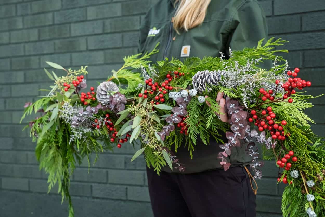 A person in a green jacket holds a festive garland with pine branches, red berries, silver pinecones, and green foliage against a dark green brick wall.