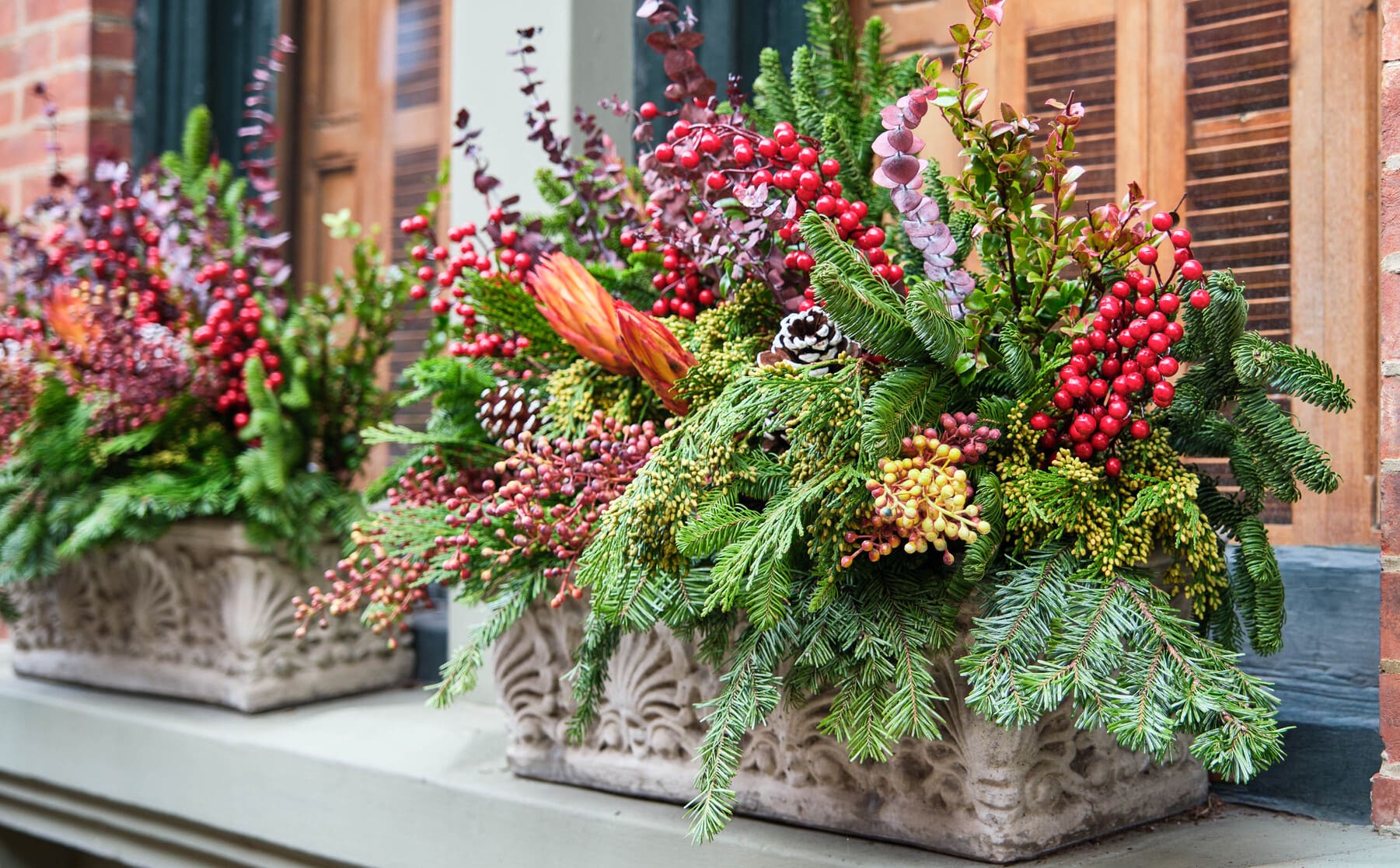 Stone planters on a porch ledge filled with evergreen branches, red berries, pinecones, and dried foliage in front of wooden shutters.