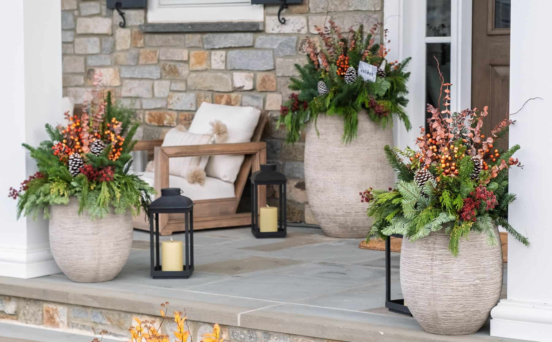 Front porch with three large planters of winter greens, berries, and pinecones, arranged beside black lanterns and a cushioned chair.