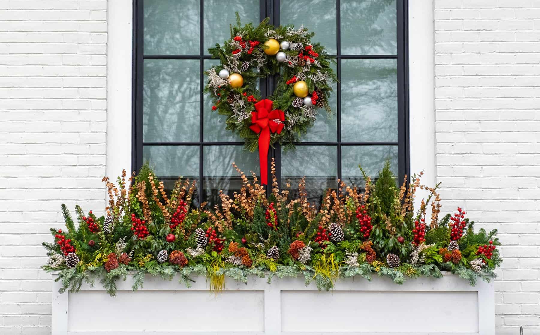 Large evergreen wreath with a red bow above a wide window box packed with fir, pinecones, and red berries on a white brick wall.