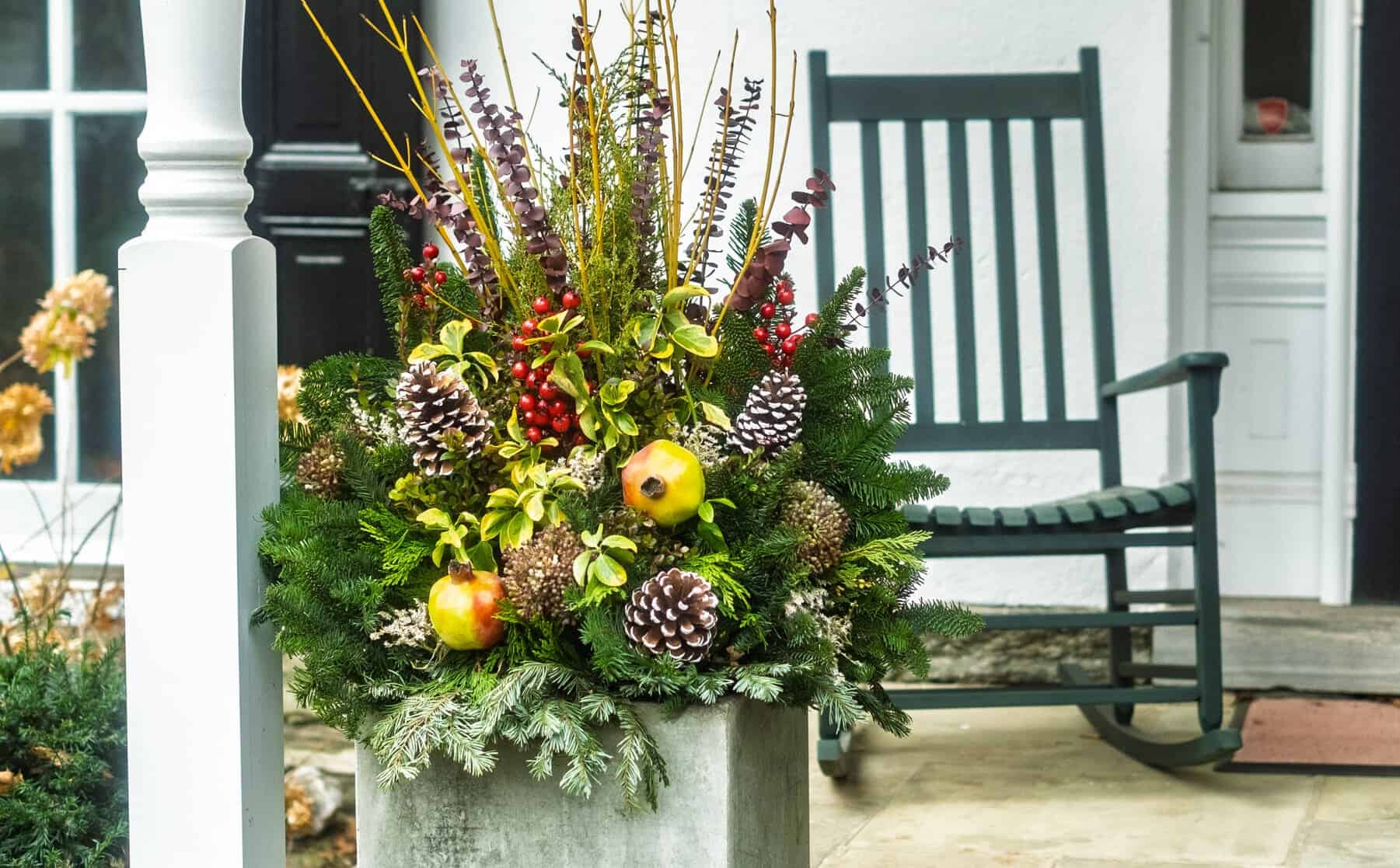 Winter planter of evergreens, pinecones, berries, and pomegranates on a porch beside a dark green rocking chair.
